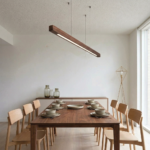 Modern wooden linear pendant light hanging above a dining table in a minimalist interior, featuring a natural wood finish and warm LED lighting.